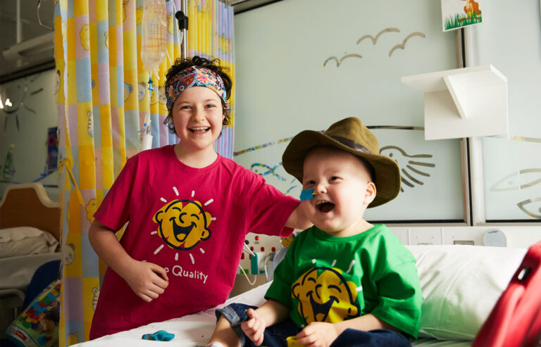 Two kids wearing Camp Quality t-shirts laugh on a hospital ward